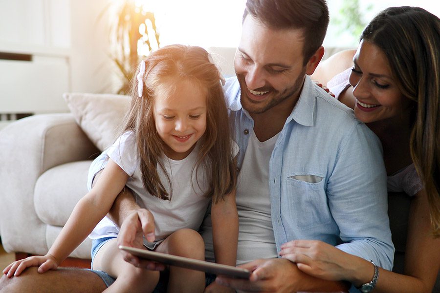 Home, Condo, Renters Quote - Father with Daught Sitting on the Living Room Floor Looking at a Tablet While Mother is Laying Behind Them on a Couch Smiling